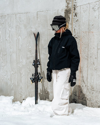 Person in winter clothing holding skis against a concrete wall with snow on the ground