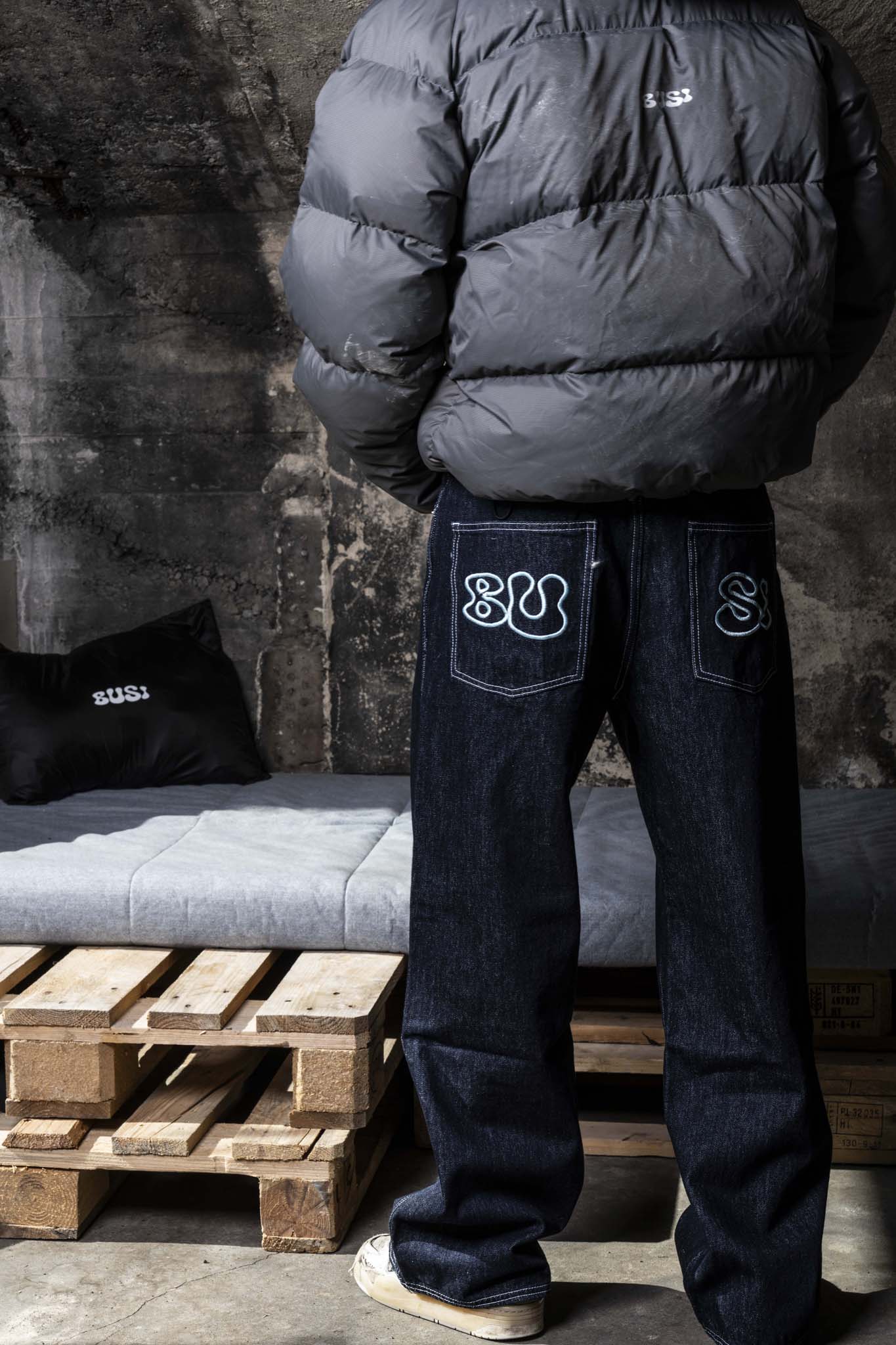 Person wearing a dark puffer jacket and jeans with visible branding, standing against a textured wall.