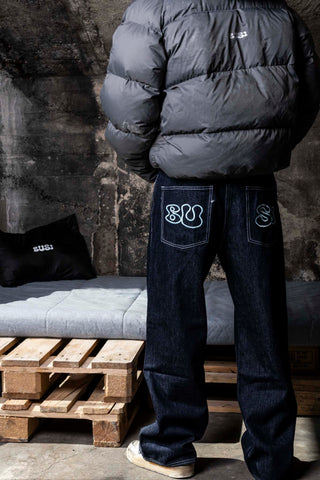 Person wearing a dark puffer jacket and jeans with visible branding, standing against a textured wall.