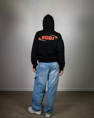 Person wearing a black hoodie with red text and blue jeans against a plain wall.