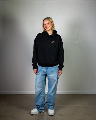 Person wearing a black hoodie and blue jeans standing against a plain white wall.