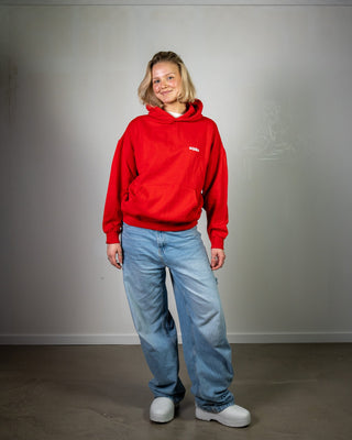 Person wearing a red hoodie and blue jeans standing against a plain wall.