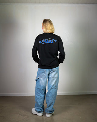 Person wearing a black sweatshirt with blue text and light blue jeans standing against a plain white wall.