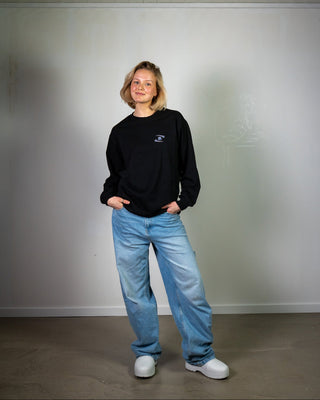 Person wearing a black sweatshirt and blue jeans standing against a plain white wall.
