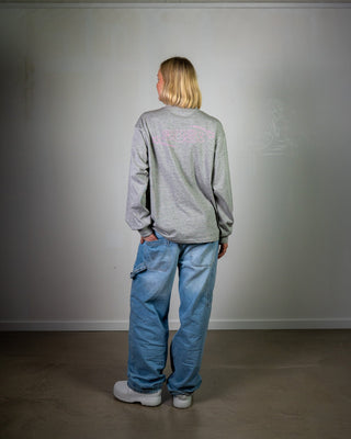 Person wearing a gray sweatshirt and blue jeans standing against a plain white wall.