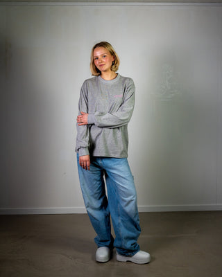 Person wearing a gray sweatshirt and blue jeans standing against a plain white wall.